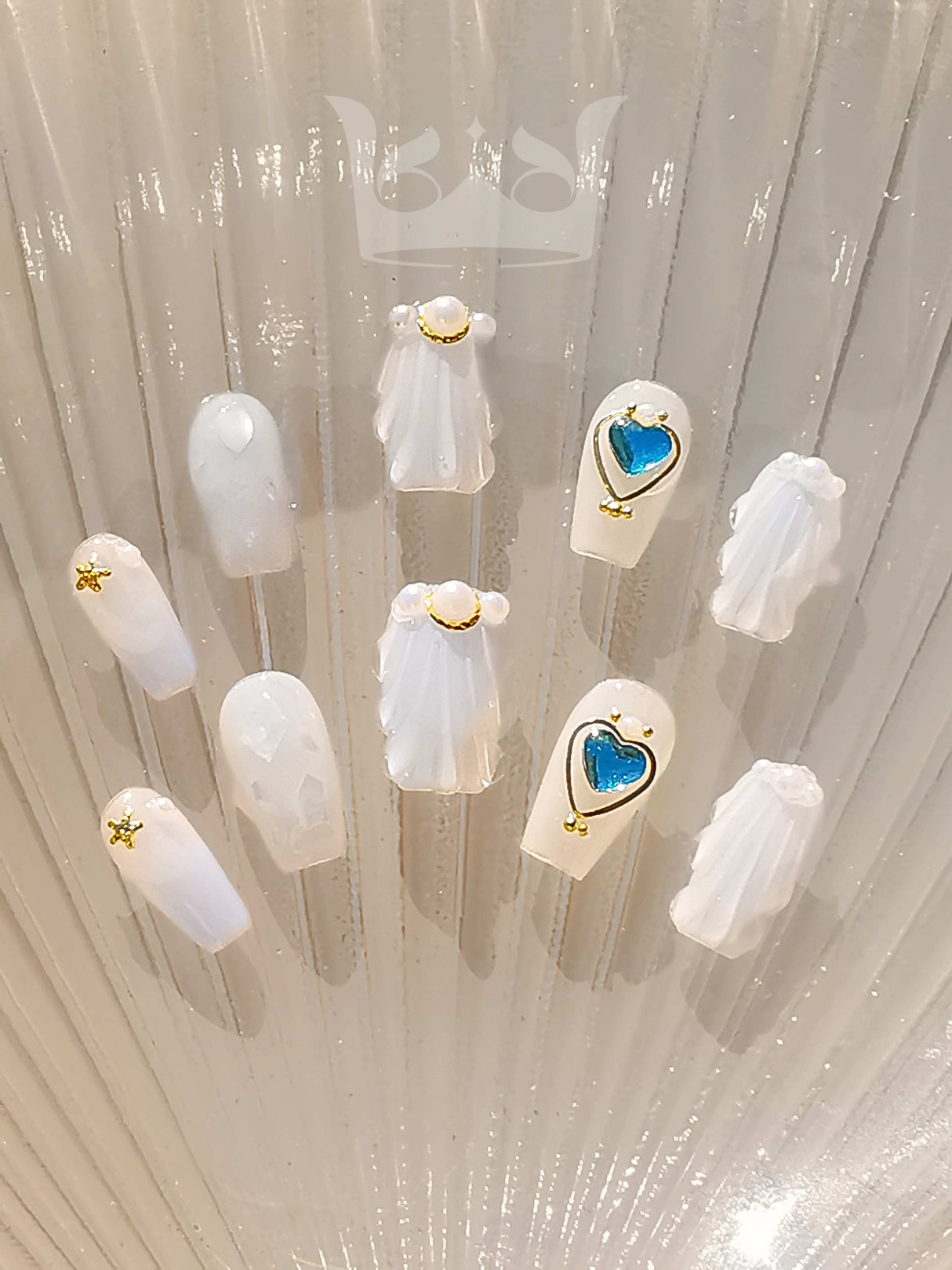 Luxury nail tips with white/translucent base, gold accents, pearls, and heart-shaped blue glittery embellishment for fancy look on special occasions.
