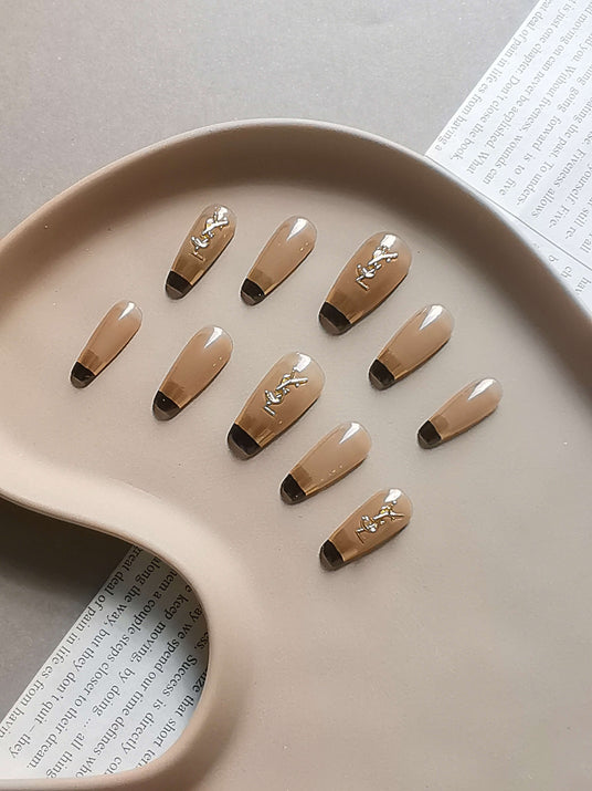 These press-on nails are designed for fashion/beauty shoots, ads, or social media posts. They feature a nude base, black tips, gold accents, and a minimalistic presentation.