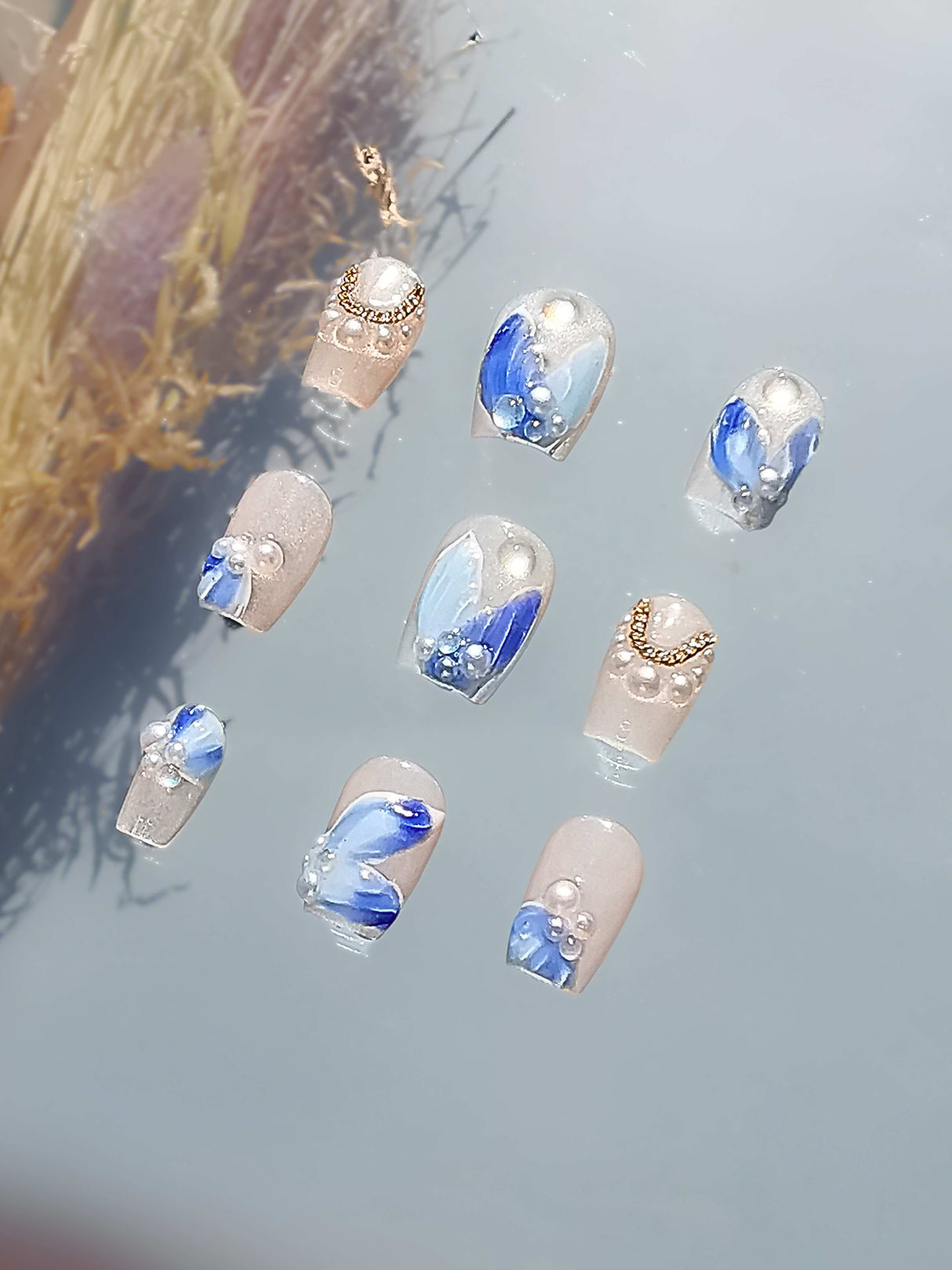 These press-on nails feature an elegant and artistic design with blue accents, rhinestones, beads, metallic elements, encapsulated design, and glossy finish.