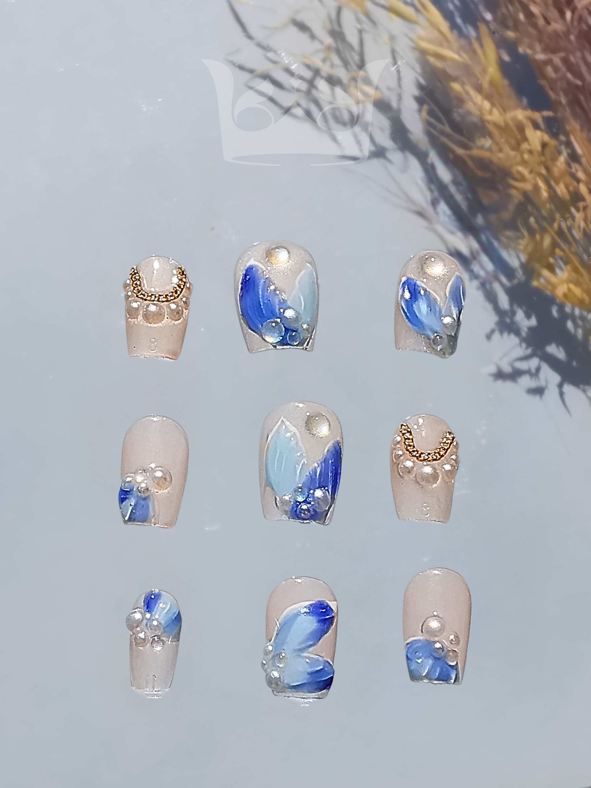 Sophisticated nails with a soft, neutral base color and blue floral accents, rhinestones, and embellishments. Glossy finish, moderate length, and varied designs.