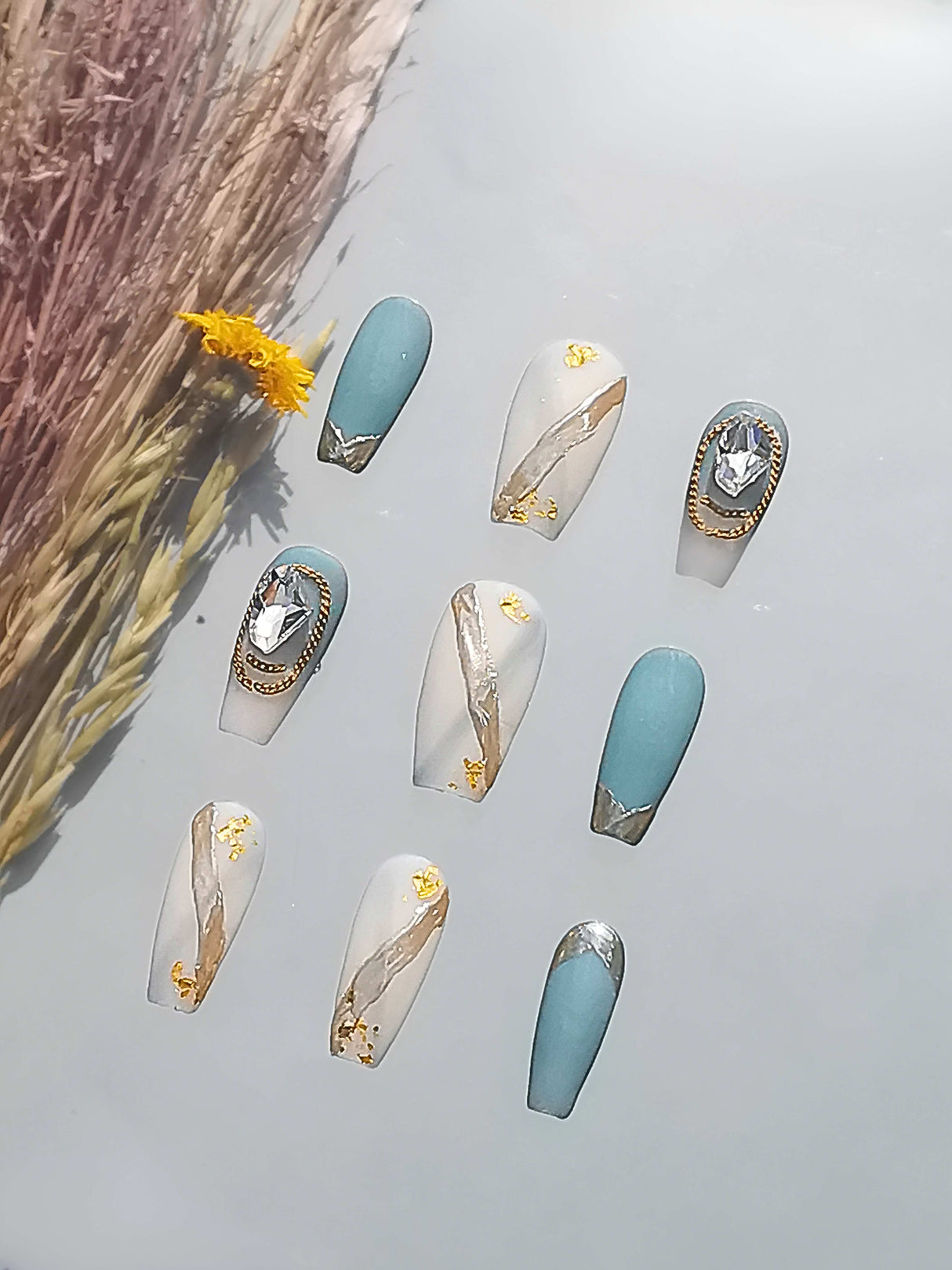 These acrylic nails with trendy designs, gold foil accents, and jeweled embellishments. The muted blue and natural tones with different finishes add a delicate aesthetic.