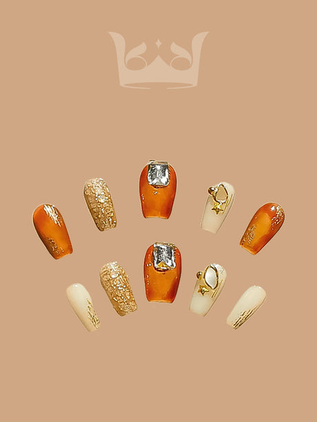 These press-on nails have shades of orange, gold, and cream, gold glitter, metallic gold foil accents, and a small gold ring or hoop embellishment.