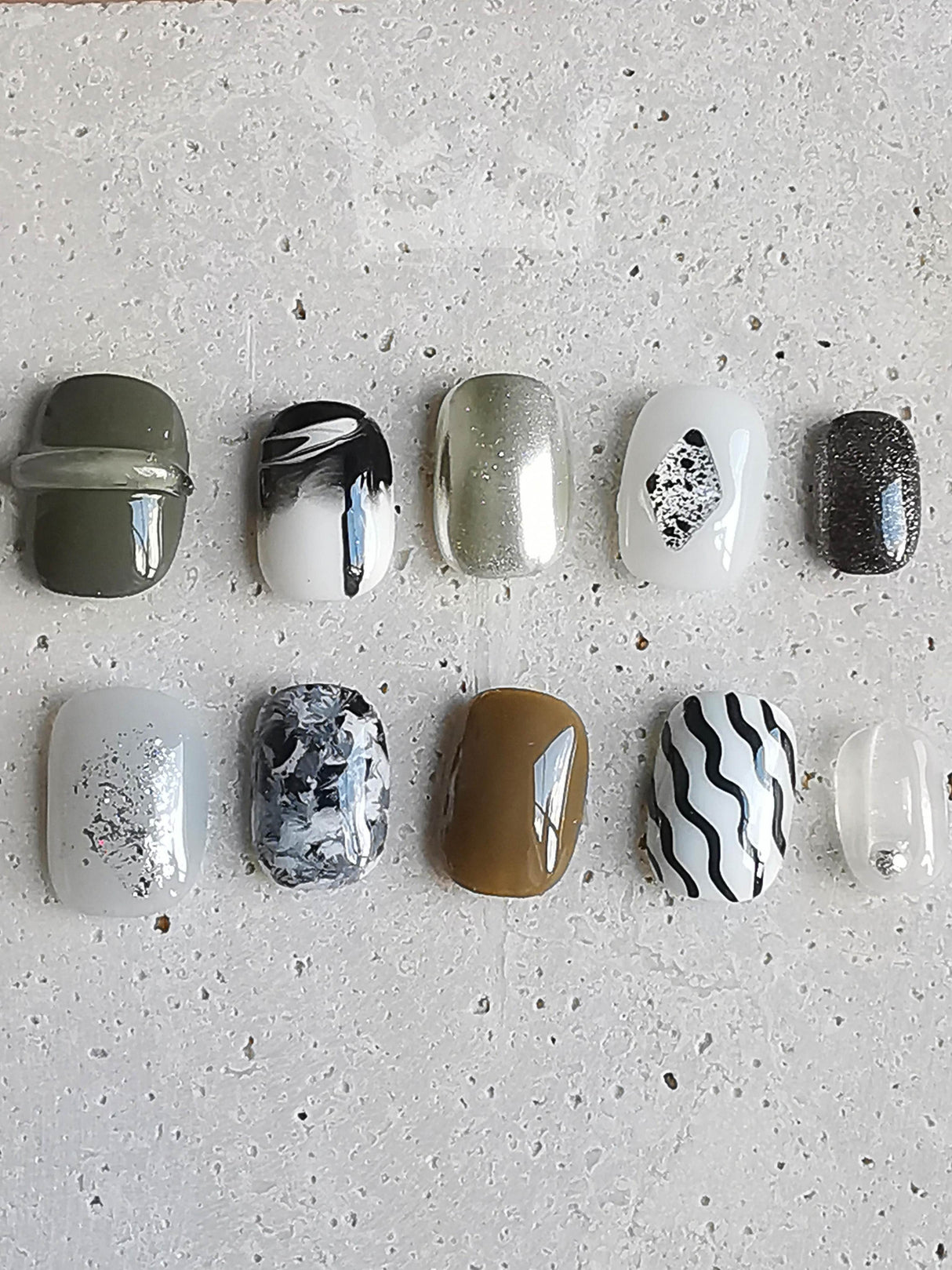 These press-on nails have glitter, stripes, marbled designs, and embellishments. The overall aesthetic is modern and stylish, with a focus on dark and neutral tones and a touch of glamour.