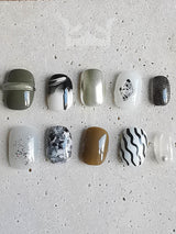 These press-on nails have glitter, stripes, marbled designs, and embellishments. The overall aesthetic is modern and stylish, with a focus on dark and neutral tones and a touch of glamour.