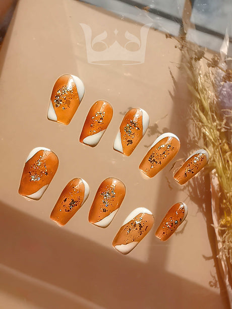 These trendy nails are painted in glossy orange with gold glitter, of varying sizes to fit different nails, and complemented by natural elements.