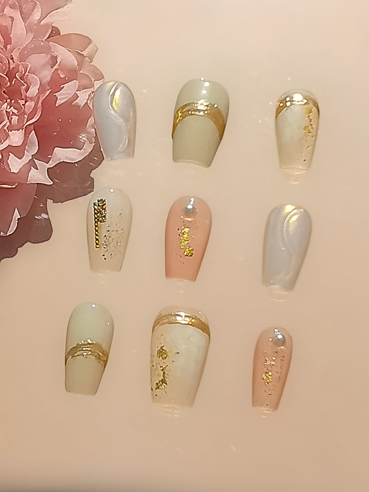 These press-on nails featuring a soft pastel color scheme, gold accents, marbled textures, 3D embellishments, and a squared-off shape with a glossy finish.