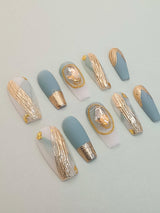 Luxurious and elegant nails with blue-gray and gold color scheme, gold foil embellishments, and vintage accents perfect for special occasions or fashion statements.