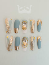 These press-on nails have a luxurious and modern design. They include a pastel blue matte finish, gold foil details, and small gold embellishments, creating an elegant and modern aesthetic.