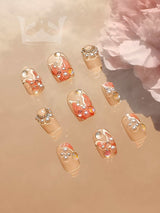 These press-on nails have a clear/nude base, glitter, rhinestones, metallic/pearlescent accents, and unique patterns, with acrylic flower.